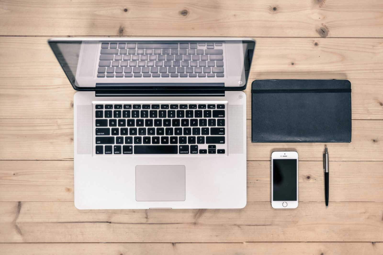 Top view of a tidy workspace featuring a laptop, smartphone, notebook, and pen on a wooden desk.