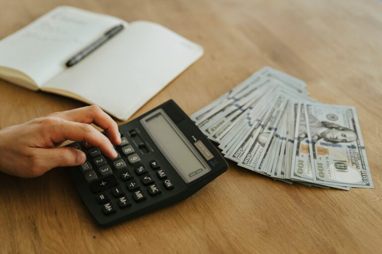 Close-up of a hand using a calculator with cash and a notebook on a wooden table.