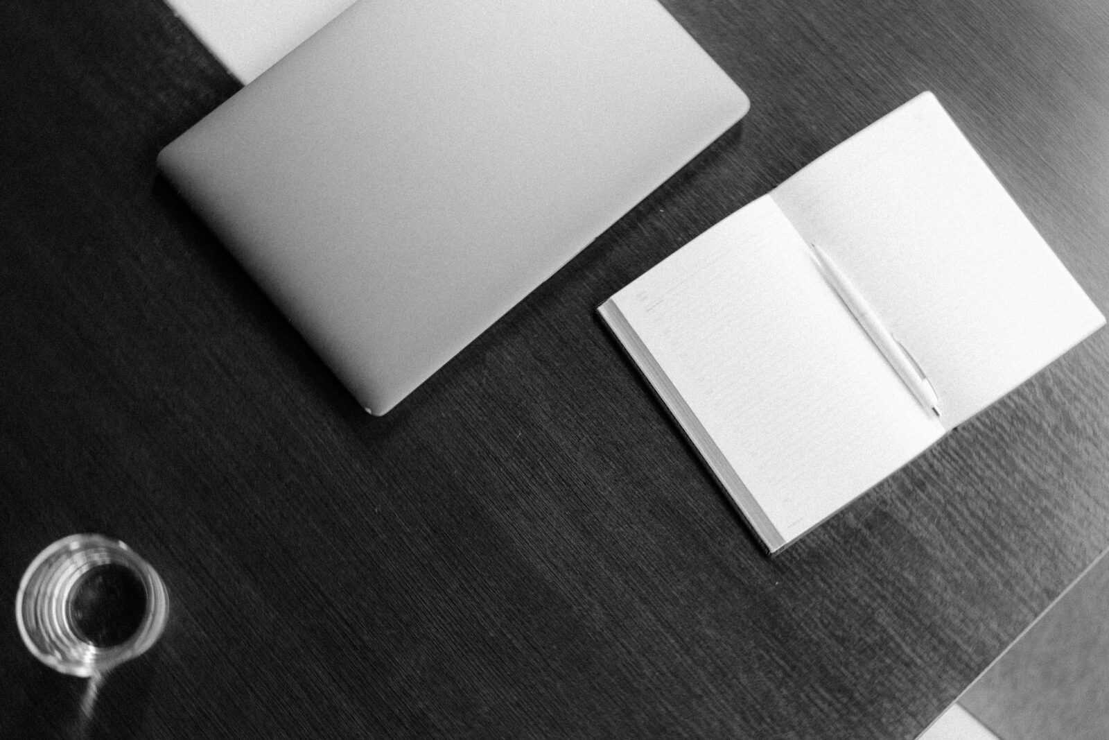 A grayscale minimalist office setup featuring a notebook, laptop, and glass on a wooden desk.